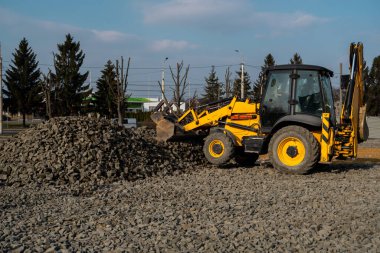 İnşaat alanında çakıllı çalışan sarı tekerlek doldurucu kazıyıcı makinesi. Asfaltlama için temel hazırlıklar yapılıyor. Yol inşaatı. Park binası..