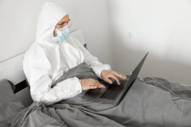 Koruyucu beyaz takım elbiseli ve tıbbi maskeli bir adam Coronavirus salgını yüzünden evde dizüstü bilgisayarla çalışıyor. Salgın sırasında uzaktan çalışma. COVID-19 karantina sırasında evde kal.
