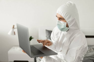 Koruyucu beyaz takım elbiseli ve tıbbi maskeli bir adam Coronavirus salgını yüzünden evde dizüstü bilgisayarla çalışıyor. Salgın sırasında uzaktan çalışma. COVID-19 karantina sırasında evde kal.