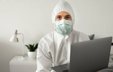 Koruyucu beyaz takım elbiseli ve tıbbi maskeli bir adam Coronavirus salgını yüzünden evde dizüstü bilgisayarla çalışıyor. Salgın sırasında uzaktan çalışma. COVID-19 karantina sırasında evde kal.
