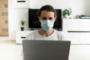 Maskeli bir adam koronavirüs salgını yüzünden evde dizüstü bilgisayarla çalışıyor. Salgın sırasında uzaktan çalışma. COVID-19 karantina sırasında evde kal. Serbest Çalışan.