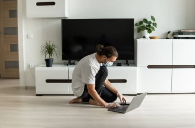 Maskeli bir adam oturma odasında bir katta oturuyor ve Coronavirus yüzünden dizüstü bilgisayarla evden çalışıyor. Salgın sırasında uzaktan çalışma. COVID-19 karantinası sırasında evde kal. Serbest Çalışan.