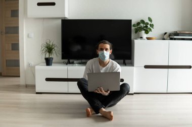Maskeli bir adam oturma odasında bir katta oturuyor ve Coronavirus yüzünden dizüstü bilgisayarla evden çalışıyor. Salgın sırasında uzaktan çalışma. COVID-19 karantinası sırasında evde kal. Serbest Çalışan.