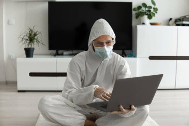 Koruyucu giysili ve tıbbi maskeli bir adam oturma odasında dizüstü bilgisayarla oturuyor çünkü Coronavirus yüzünden. Salgın sırasında uzaktan çalışma. COVID-19 karantinası sırasında evde kal. Serbest Çalışan.