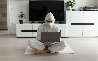 Koruyucu giysili ve tıbbi maskeli bir adam oturma odasında dizüstü bilgisayarla oturuyor çünkü Coronavirus yüzünden. Salgın sırasında uzaktan çalışma. COVID-19 karantinası sırasında evde kal. Serbest Çalışan.