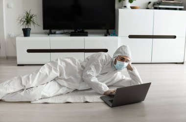 Koruyucu giysili ve tıbbi maskeli bir adam, Coronavirus yüzünden oturma odasında dizüstü bilgisayarla yerde yatıyor. Salgın sırasında uzaktan çalışma. COVID-19 karantinası sırasında evde kal. Serbest Çalışan.