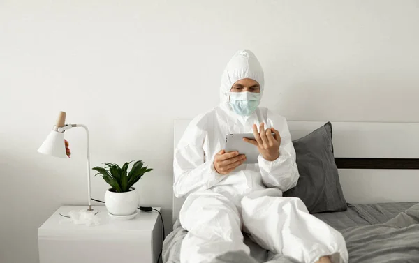 Man in protective white suit and medical mask is working from home on a bed with a tablet because of coronavirus epidemic. Remote work during pandemic. Stay home during COVID-19 quarantine concept.