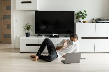 Tıbbi maskeli bir adam oturma odasında yerde yatıyor ve evde dizüstü bilgisayarla çalışıyor ve Coronavirüs salgını nedeniyle web kamerasıyla arkadaşlarına ya da iş ortaklarına merhaba diyor..