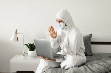 Koruyucu beyaz takım elbiseli ve tıbbi maskeli bir adam evde dizüstü bilgisayarla çalışıyor ve Coronavirüs salgını yüzünden arkadaşlarına ya da iş ortaklarına merhaba diyor..