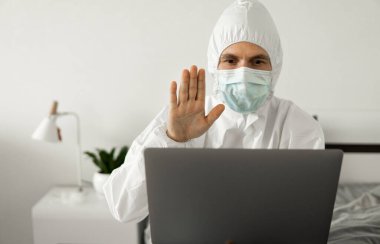 Koruyucu beyaz takım elbiseli ve tıbbi maskeli bir adam evde dizüstü bilgisayarla çalışıyor ve Coronavirüs salgını yüzünden arkadaşlarına ya da iş ortaklarına merhaba diyor..