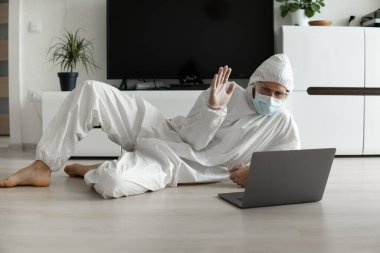 Koruyucu giysili ve tıbbi maskeli bir adam oturma odasında dizüstü bilgisayarla yatıyor ve Coronavirüs salgını nedeniyle web kamerasıyla arkadaşlarına ya da iş arkadaşlarına merhaba diyor..