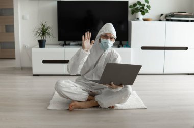 Koruyucu giysili ve tıbbi maskeli bir adam oturma odasında dizüstü bilgisayarla oturuyor çünkü Coronavirus yüzünden. Salgın sırasında uzaktan çalışma. COVID-19 karantinası sırasında evde kal. Serbest Çalışan.