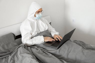 Koruyucu beyaz takım elbiseli ve tıbbi maskeli bir adam Coronavirus salgını yüzünden evde dizüstü bilgisayarla çalışıyor. Salgın sırasında uzaktan çalışma. COVID-19 karantina sırasında evde kal.