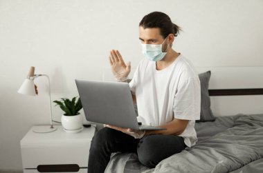 Tıbbi maskeli bir adam evde dizüstü bilgisayarla çalışıyor ve Coronavirus yüzünden arkadaşlarıyla veya iş ortaklarıyla web kamerasıyla konuşuyor..