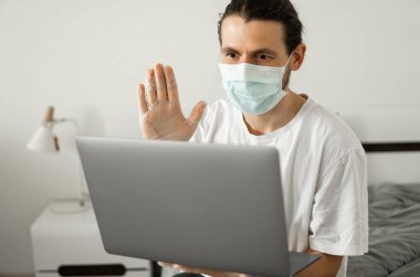 Tıbbi maskeli bir adam evde dizüstü bilgisayarla çalışıyor ve Coronavirus yüzünden arkadaşlarıyla veya iş ortaklarıyla web kamerasıyla konuşuyor..