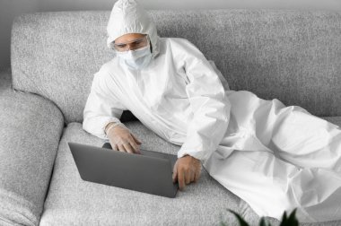 Koruyucu takım elbiseli ve tıbbi maskeli bir adam oturma odasındaki kanepede dizüstü bilgisayarla yatıyor çünkü koronavirüs var. Salgın sırasında uzaktan çalışma. COVID-19 karantinası sırasında evde kal. Serbest Çalışan.