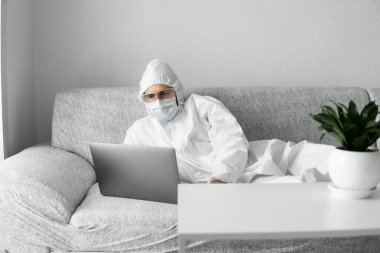Koruyucu takım elbiseli ve tıbbi maskeli bir adam oturma odasındaki kanepede dizüstü bilgisayarla yatıyor çünkü koronavirüs var. Salgın sırasında uzaktan çalışma. COVID-19 karantinası sırasında evde kal. Serbest Çalışan.