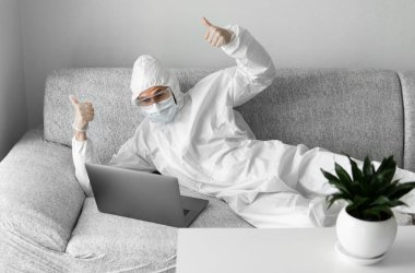 Koruyucu takım elbiseli ve tıbbi maskeli bir adam oturma odasındaki kanepede dizüstü bilgisayarla yatıyor ve projeden mutlu olduğu için iki parmağını kaldırıyor. Salgın COVID-19 karantinasında uzaktan çalışma.