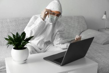 Koruyucu beyaz takım elbiseli ve tıbbi maskeli bir adam Coronavirus salgını nedeniyle evinde dizüstü bilgisayarı ve tableti olan bir koltukta çalışıyor. Salgın sırasında uzaktan çalışma. COVID-19 karantinası sırasında evde kal