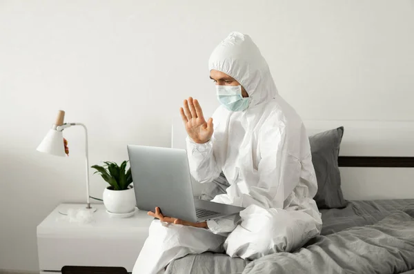 Man in protective white suit and medical mask is working from home on a bed with laptop and showing hello to his friends or business partners because of coronavirus epidemic.