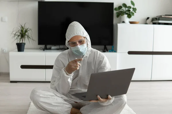 Man in protective suit and medical mask is sitting on a floor in his living room with a laptop because of coronavirus. Remote work during pandemic. Stay home during COVID-19 quarantine. Freelancer.