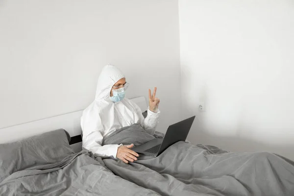 El hombre con traje blanco protector y máscara médica está trabajando desde casa en una cama con computadora portátil debido a la epidemia de coronavirus. Trabajo remoto durante una pandemia. Permanecer en casa durante el concepto de cuarentena COVID-19 . —  Fotos de Stock