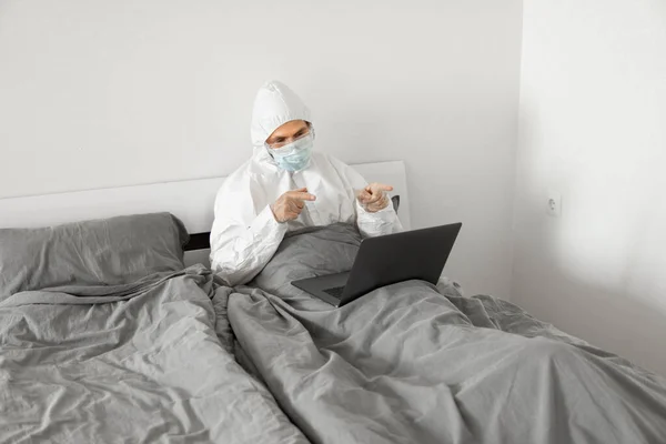 Man in protective white suit and medical mask is working from home in a bed with laptop because of coronavirus epidemic. Remote work during pandemic. Stay home during COVID-19 quarantine concept.