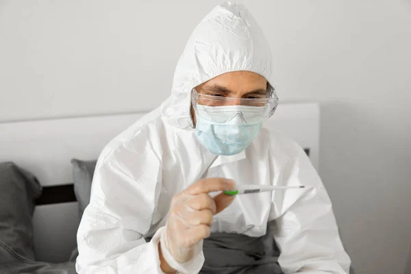 Man in white protective suit, medical mask, glasses and rubber gloves is holding in hand thermometer in a bed at his home. Epidemic, pandemic, coronavirus, 2019-ncov, covid-19, virus.