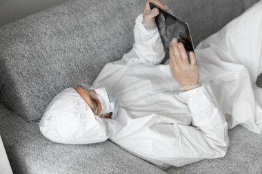 Koruyucu beyaz takım elbiseli ve tıbbi maskeli bir adam Coronavirüs salgını yüzünden elinde tabletle evde yatıyor. Salgın sırasında uzaktan çalışma. COVID-19 karantinası sırasında evde kal