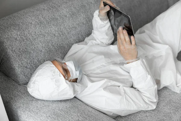Man in protective white suit and medical mask is working from home lying on a sofa with a tablet because of coronavirus epidemic. Remote work during pandemic. Stay home during COVID-19 quarantine