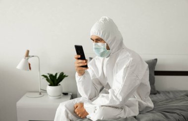 Koruyucu beyaz takım elbiseli ve tıbbi maskeli bir adam Coronavirüs salgını yüzünden evinde yatağında oturan bir telefonu kullanıyor. Salgın sırasında uzaktan çalışma. COVID-19 karantina sırasında evde kal.