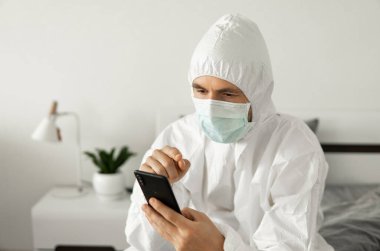 Koruyucu beyaz takım elbiseli ve tıbbi maskeli bir adam Coronavirüs salgını yüzünden evinde yatağında oturan bir telefonu kullanıyor. Salgın sırasında uzaktan çalışma. COVID-19 karantina sırasında evde kal.