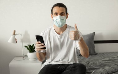 Maskeli bir adam evinde telefon kullanıyor. Coronavirüs salgını yüzünden yatağın üzerinde oturuyor. Salgın sırasında uzaktan çalışma. COVID-19 karantina sırasında evde kal.
