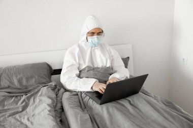 Koruyucu beyaz takım elbiseli ve tıbbi maskeli bir adam evde dizüstü bilgisayarlı bir yatakta çalışıyor ve web kamerasıyla arkadaşı veya iş arkadaşlarıyla konuşuyor. Coronavirüs salgını. Salgın sırasında uzak çalışma.