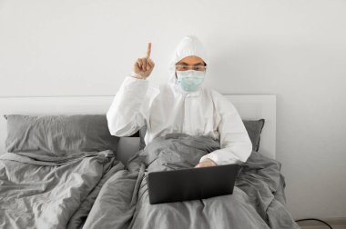 Koruyucu beyaz takım elbiseli ve tıbbi maskeli bir adam evde dizüstü bilgisayarlı bir yatakta çalışıyor ve web kamerasıyla arkadaşı veya iş arkadaşlarıyla konuşuyor. Coronavirüs salgını. Salgın sırasında uzak çalışma.