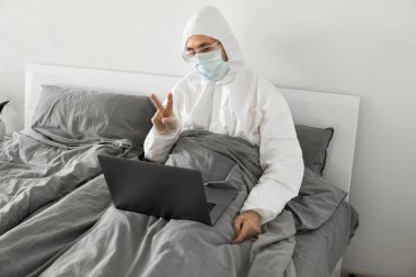 Koruyucu beyaz takım elbiseli ve tıbbi maskeli bir adam evde dizüstü bilgisayarlı bir yatakta çalışıyor ve web kamerasıyla arkadaşı veya iş arkadaşlarıyla konuşuyor. Coronavirüs salgını. Salgın sırasında uzak çalışma.