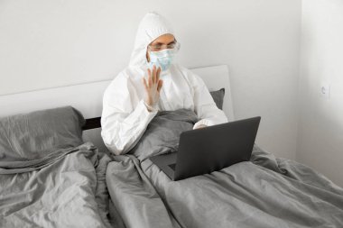 Koruyucu beyaz takım elbiseli ve tıbbi maskeli bir adam evde dizüstü bilgisayarlı bir yatakta çalışıyor ve web kamerasıyla arkadaşı veya iş arkadaşlarıyla konuşuyor. Coronavirüs salgını. Salgın sırasında uzak çalışma.