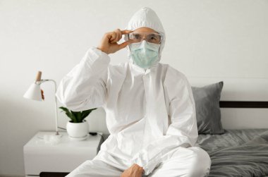 Beyaz koruyucu takım elbiseli, tıbbi maskeli ve lastik eldivenli adamlar yatak odasındaki bir yatakta otururken koronavirüs salgını tehdidi altında. Salgın, Coronavirus covid 19 salgını. Solunum cihazındaki adam..