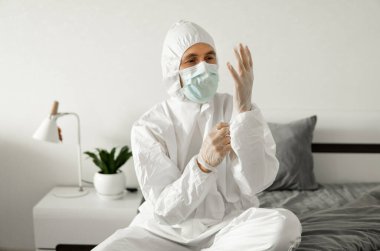 Beyaz koruyucu takım elbiseli, tıbbi maskeli ve lastik eldivenli adamlar yatak odasındaki bir yatakta otururken koronavirüs salgını tehdidi altında. Salgın, Coronavirus covid 19 salgını. Solunum cihazındaki adam..