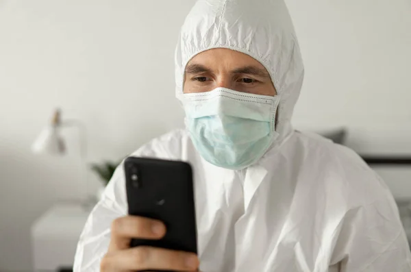 Man in protective white suit and medical mask is using a phone at his home sitting on a bed because of coronavirus epidemic. Remote work during pandemic. Stay home during COVID-19 quarantine concept.