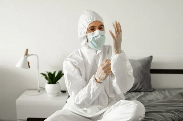 Men in white protective suit, medical mask and rubber gloves is sitting on a bed in his bedroom while coronavirus pandemic threat. Epidemic, pandemic of coronavirus covid 19. Man in respirator.