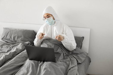 Koruyucu beyaz takım elbiseli ve tıbbi maskeli bir adam evde dizüstü bilgisayarlı bir yatakta çalışıyor ve web kamerasıyla arkadaşı veya iş arkadaşlarıyla konuşuyor. Coronavirüs salgını. Salgın sırasında uzak çalışma.