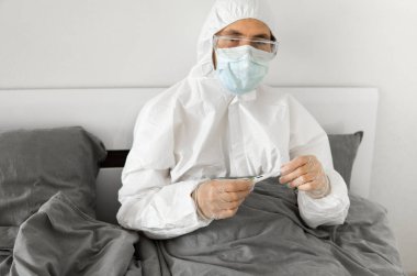 Beyaz koruyucu takım elbiseli, tıbbi maskeli, gözlüklü ve lastik eldivenli bir adam evinde bir yatakta el termometresi tutuyor. Salgın, salgın, koronavirüs, 2019-ncov, covid-19, virüs.