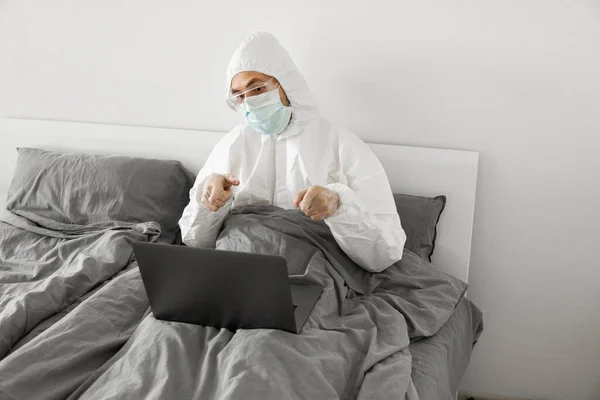 Koruyucu beyaz takım elbiseli ve tıbbi maskeli bir adam evde dizüstü bilgisayarlı bir yatakta çalışıyor ve web kamerasıyla arkadaşı veya iş arkadaşlarıyla konuşuyor. Coronavirüs salgını. Salgın sırasında uzak çalışma. — Stok fotoğraf