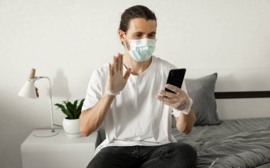 Tıbbi maskeli bir adam evinde bir telefon kullanıyor bir yatakta oturuyor ve web kamerasıyla arkadaşına ya da iş arkadaşına merhaba diyor. Coronavirüs salgını. Salgın sırasında uzak çalışma.