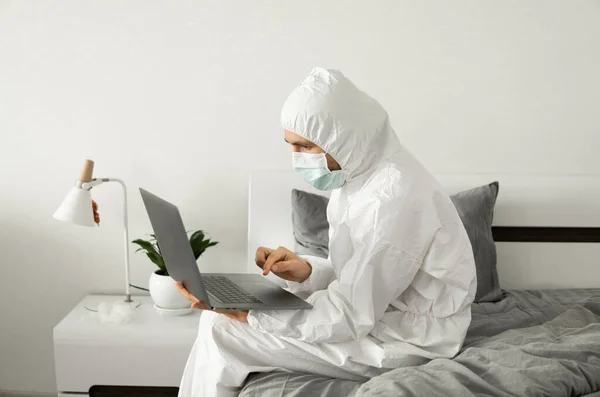 Man in protective white suit and medical mask is working from home on a bed with laptop because of coronavirus epidemic. Remote work during pandemic. Stay home during COVID-19 quarantine concept.