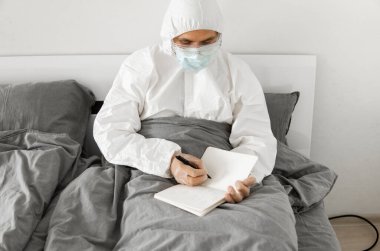Korumacı beyaz takım elbiseli ve yüz maskeli bir adamın yatak odasında oturmasının nedeni 2019 yılının defterinde bir gün sayıp koronavirüs karantinasının sona ermesini beklemesi..