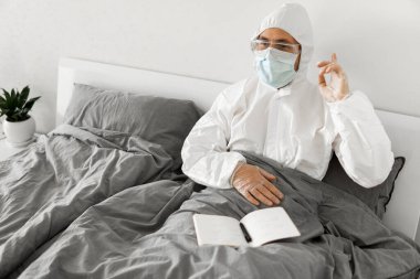 Korumacı beyaz takım elbiseli ve yüz maskeli bir adamın yatak odasında oturmasının nedeni 2019 yılının defterinde bir gün sayıp koronavirüs karantinasının sona ermesini beklemesi..