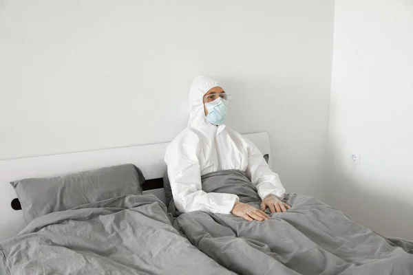 Retrato del hombre en traje blanco protector y máscara facial sentado en su cama en su dormitorio debido a covid-2019 y esperar a que finalice la cuarentena del coronavirus. Autoaislamiento . —  Fotos de Stock