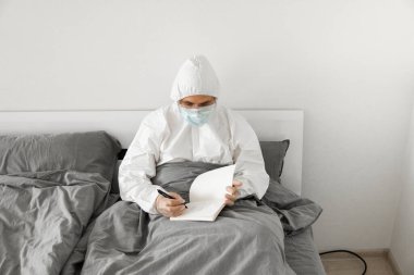 Korumacı beyaz takım elbiseli ve yüz maskeli bir adamın yatak odasında oturmasının nedeni 2019 yılının defterinde bir gün sayıp koronavirüs karantinasının sona ermesini beklemesi..